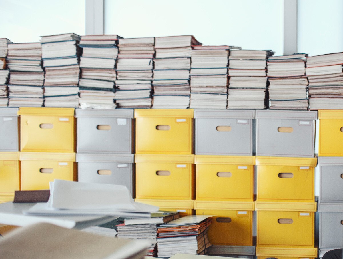 Cluttered office workspace with stacks of discovery documents in yellow and gray boxes, showing a busy work environment